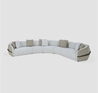 A large, modular, curved sectional sofa upholstered in a textured, light-colored fabric with a subtle speckled pattern. The sofa features several individual seating modules forming a gentle arc. It is accompanied by throw pillows in complementary neutral tones. Distinctive design elements include curved armrests and side panels with inset wood-look trim, adding a contemporary and sophisticated aesthetic. — 3D model