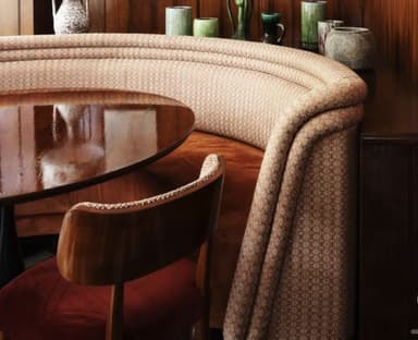 A custom-designed curved banquette seating unit, upholstered in a light-colored fabric with a subtle geometric pattern. It features a channel-tufted backrest and appears to be built into a corner or against a wall, forming a cozy dining or lounge area with a round, highly polished dark wood table and a wooden dining chair with an upholstered seat. — 3D model