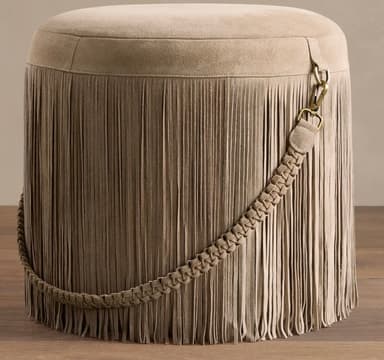 A cylindrical upholstered ottoman featuring a full skirt of cascading fringe around its perimeter. The piece is covered in a soft, matte taupe or greige suede-like material. A decorative braided strap, also in the same material, wraps diagonally around the body of the ottoman, secured by an antiqued brass-tone clasp. The top surface is flat and plush, indicating suitability for seating or footrest use. The overall aesthetic leans towards a bohemian or eclectic style. — 3D model