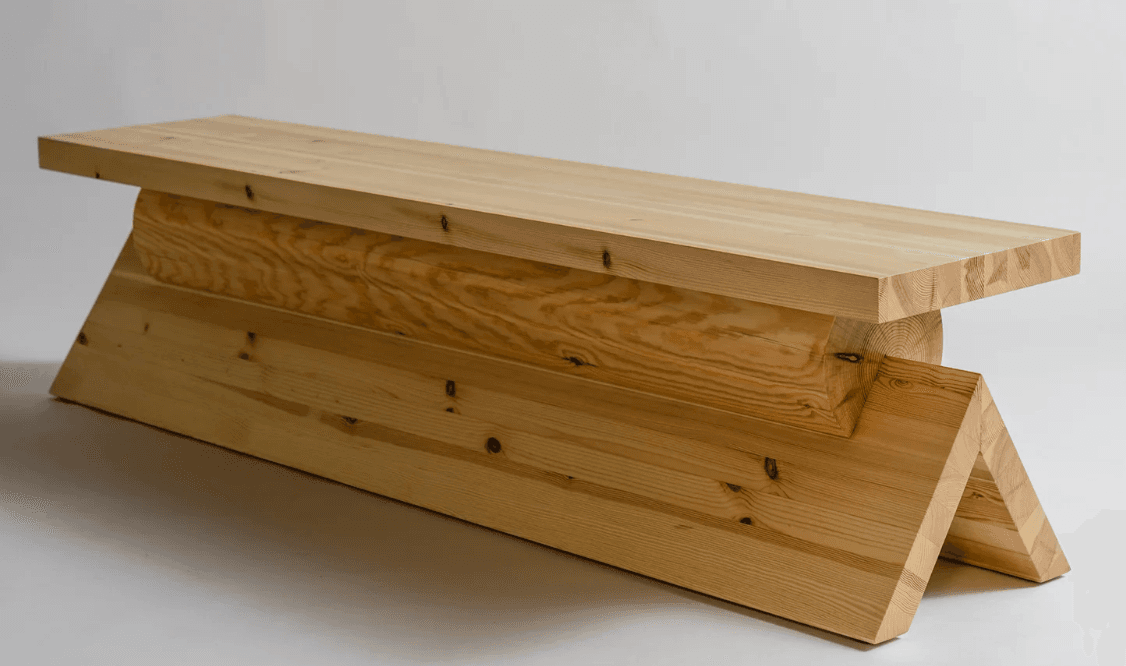 A long, low bench crafted from light-toned solid wood. It features a thick, rectangular top surface supported by a geometrically constructed base made of interlocking wooden planks or beams, presenting a robust and modern aesthetic. The visible wood grain and knots suggest a natural finish. — 3D model