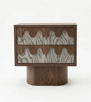A contemporary nightstand featuring two drawers with a distinctive wave-like carved wooden facade. The main body and base are constructed from dark wood, while the drawer fronts exhibit a contrasting lighter wood with prominent grain patterns, also featuring the wave motif. The piece is supported by a solid, cylindrical wooden base. — 3D model