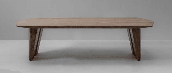 A low-profile rectangular coffee table crafted from solid wood, featuring a light brown finish that highlights its natural grain. The tabletop has subtly tapered edges, giving it a refined profile. It is supported by two sets of angular, sculptural legs that are thick and robust, contributing to a modern and sturdy aesthetic. The overall design emphasizes clean lines and a minimalist form. — 3D model
