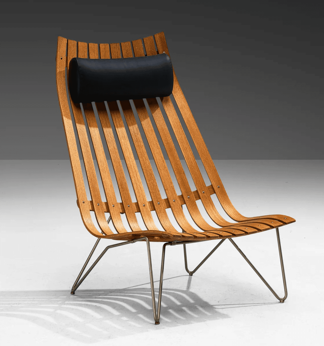 A mid-century modern lounge chair featuring a distinctive slatted wooden back and seat, supported by a minimalist metal wire frame. The chair includes a cylindrical black leather or vinyl headrest for comfort. Its design emphasizes ergonomic curves and a lightweight aesthetic. — 3D model
