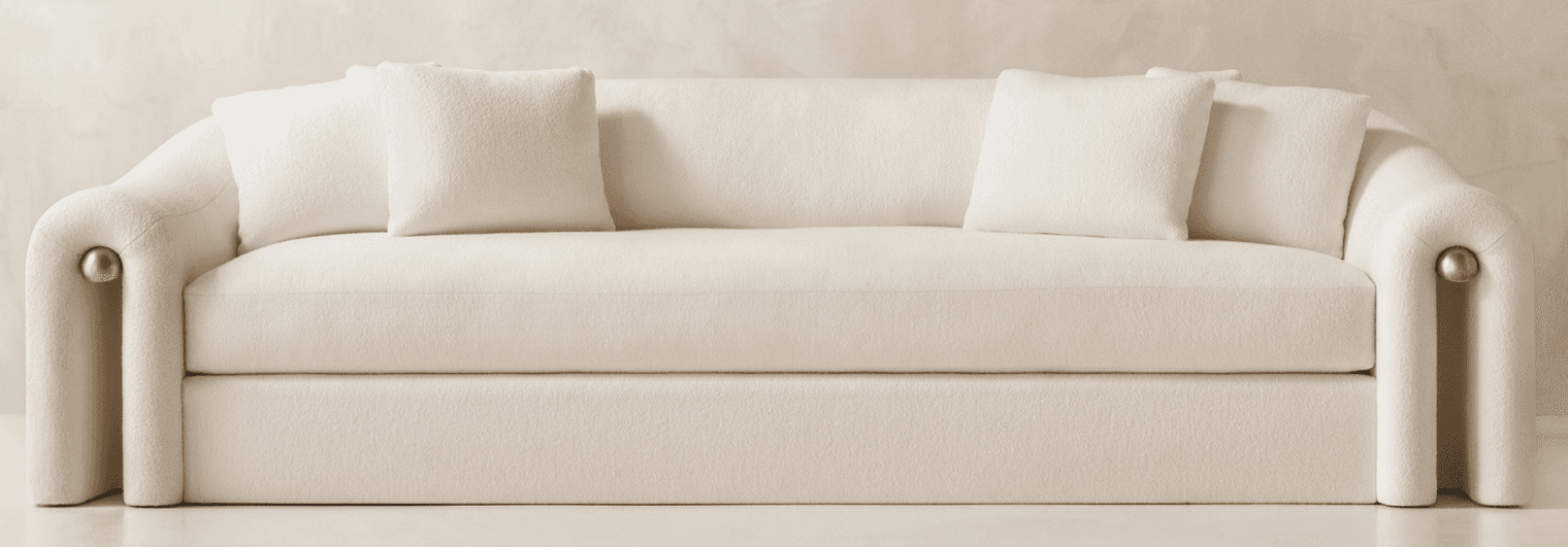 A modern three-seater sofa featuring a distinctive curved silhouette and rolled arms, upholstered in a cream-colored fabric. The arms are accented with subtle brass details at the front base. It has a single, long seat cushion and two loose back cushions. — 3D model