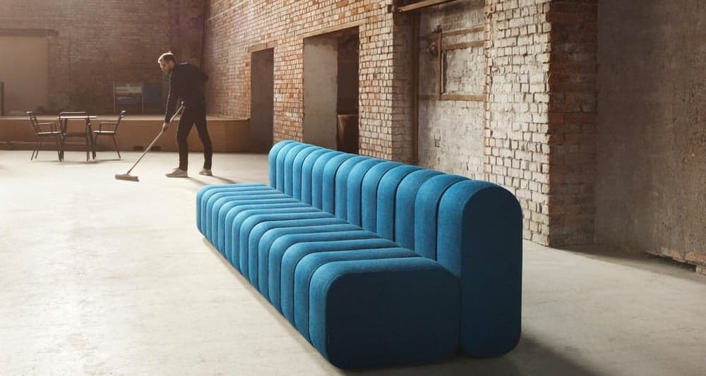 A low-profile, modular sofa with a distinctive channel-tufted design. The sofa is upholstered in a textured blue fabric and features rounded edges. It appears to be composed of two connected seating modules, forming an 'L' or angular shape when viewed from above, though the render suggests a linear arrangement with an integrated backrest. The minimalist aesthetic suggests a contemporary or modern design. — 3D model