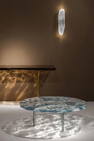 A round coffee table with a unique top designed to mimic the appearance of rippling water. The surface has an undulating texture, creating patterns of light and shadow on the floor beneath. It is supported by slender, dark-colored legs. — 3D model