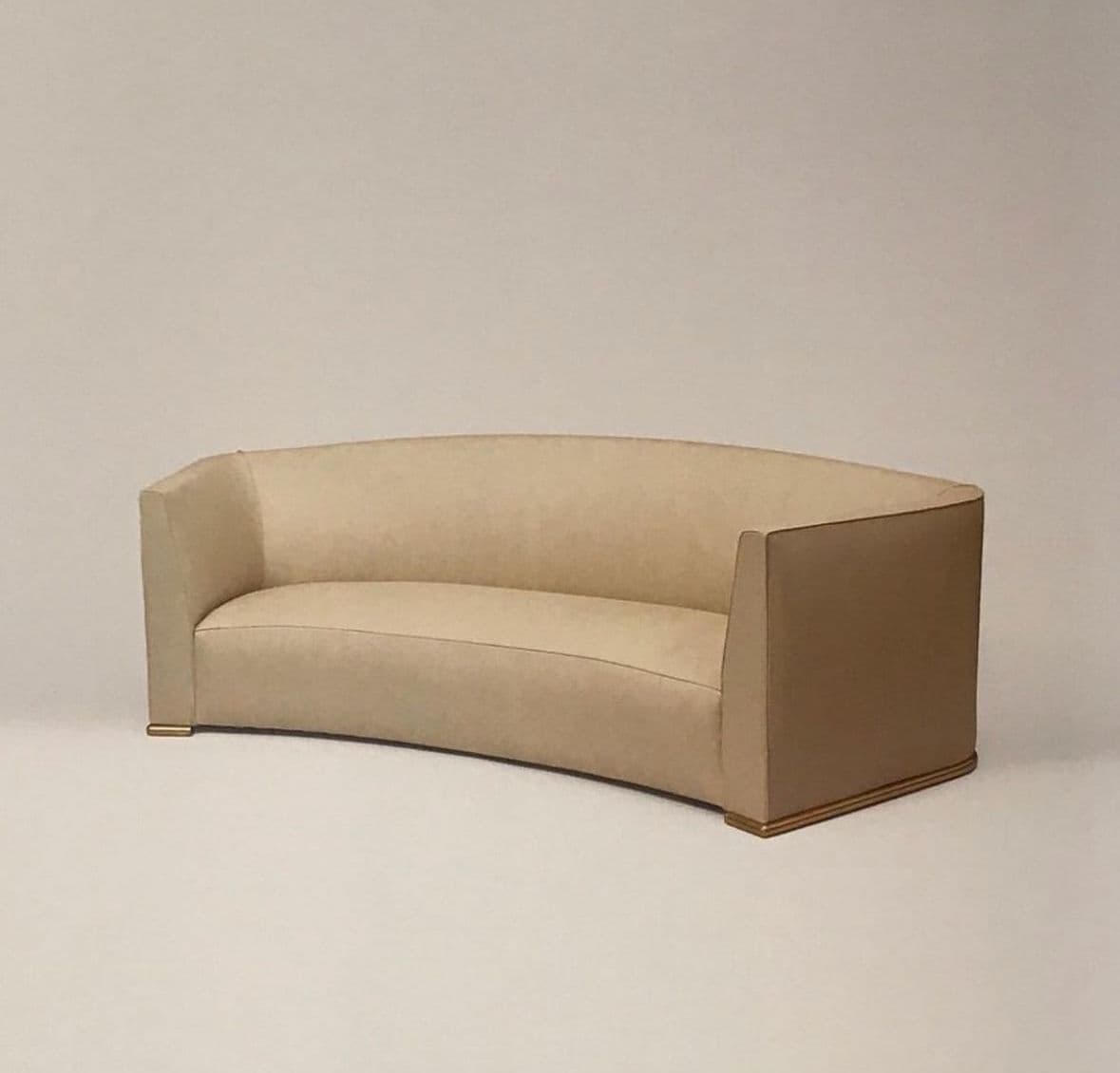 A contemporary curved sofa upholstered in light beige fabric, featuring a continuous back and armrest design, and a recessed plinth base accented with a gold-toned trim. — 3D model