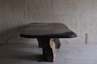 A large dining table with an irregular, organic-shaped top and two substantial, textured bases that resemble natural rock formations or tree trunks. The tabletop appears to be made of multiple panels, possibly wood or stone, with a dark, possibly metal or resin, edge banding. The bases are dark brown and have a rough, sculpted texture, giving the impression of raw, natural elements. — 3D model