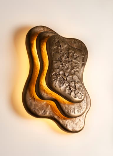 A sculptural wall sconce featuring an organic, multi-layered design in a bronze finish. The light source is positioned behind the contoured metal plates, casting a warm, indirect glow that highlights the irregular shapes and textured surfaces. The outermost layer has a rough, craggy texture, while inner layers are smoother, creating depth and visual interest. — 3D model