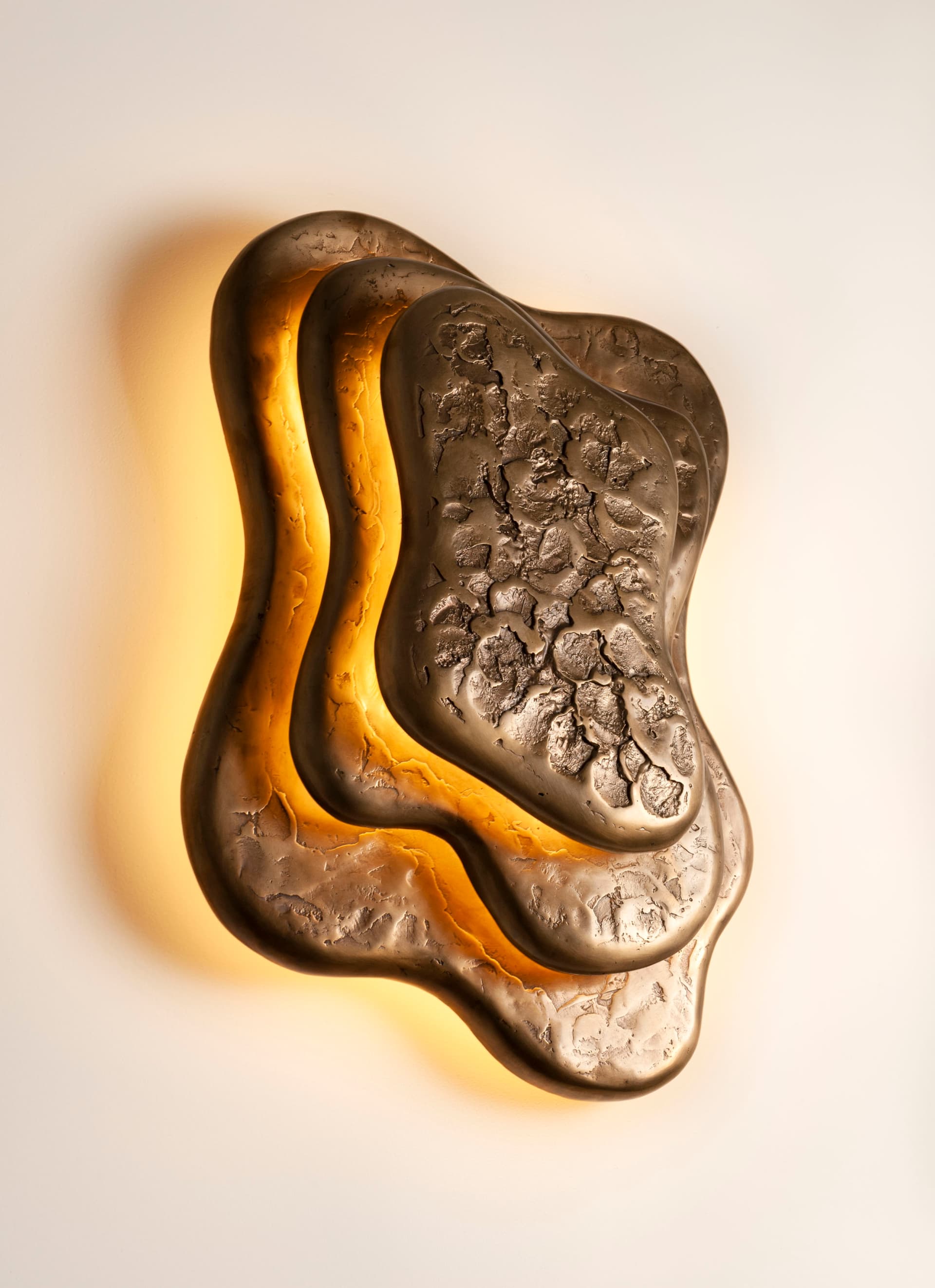 A sculptural wall sconce featuring an organic, multi-layered design in a bronze finish. The light source is positioned behind the contoured metal plates, casting a warm, indirect glow that highlights the irregular shapes and textured surfaces. The outermost layer has a rough, craggy texture, while inner layers are smoother, creating depth and visual interest. — 3D model