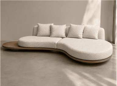 An overhead view of a contemporary, asymmetrical sectional sofa featuring a curved seating arrangement and an integrated wooden platform. The sofa is upholstered in a textured, light-colored fabric, likely bouclé, and includes several loose back cushions. The base of the sofa is connected to an organic-shaped wooden platform that extends beyond the seating modules, acting as a functional side table or display surface. The design emphasizes fluidity and modularity with soft, rounded edges. — 3D model