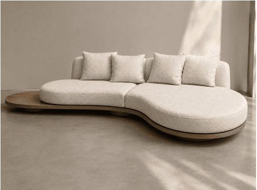 An overhead view of a contemporary, asymmetrical sectional sofa featuring a curved seating arrangement and an integrated wooden platform. The sofa is upholstered in a textured, light-colored fabric, likely bouclé, and includes several loose back cushions. The base of the sofa is connected to an organic-shaped wooden platform that extends beyond the seating modules, acting as a functional side table or display surface. The design emphasizes fluidity and modularity with soft, rounded edges. — 3D model