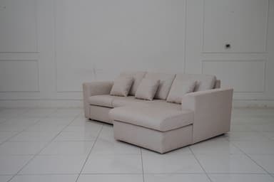 A sectional sofa with a chaise lounge extension, upholstered in a light-colored fabric with a subtle pattern. The sofa features three seat cushions and a chaise on the right side. It includes four matching throw pillows. The sofa has a modern, minimalist design with clean lines and low-profile arms and back. — 3D model
