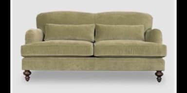 A traditional two-seater loveseat upholstered in a soft, sage green fabric, likely velvet or velour. It features rolled arms, a slightly flared backrest, and two loose back cushions. The base is supported by four dark, turned wooden feet. — 3D model