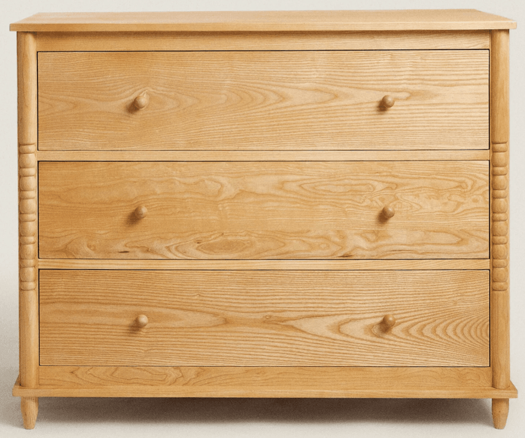 A light natural oak wood dresser with three full-width drawers, featuring simple round wood knob pulls. The dresser is characterized by its turned and fluted vertical posts at the front corners, which extend down to form short legs. The wood displays a prominent grain pattern. — 3D model