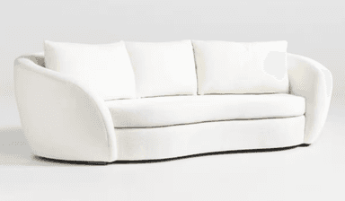 A close-up view of a cream-colored sofa, focusing on the armrest and seat cushions. The upholstery appears to be a textured boucle or similar woven fabric. The armrest has a rounded, curved profile, and the seat cushions are plush with visible stitching. The base of the sofa, where it meets the floor, is partially visible and appears to be a dark wood. — 3D model