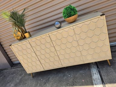 A long, rectangular sideboard featuring a contemporary design with a repetitive geometric honeycomb pattern across its four cabinet doors. The main body is a neutral cream or beige color, complemented by slender, angled brass-toned metal legs and matching brass corner accents on the top surface. — 3D model