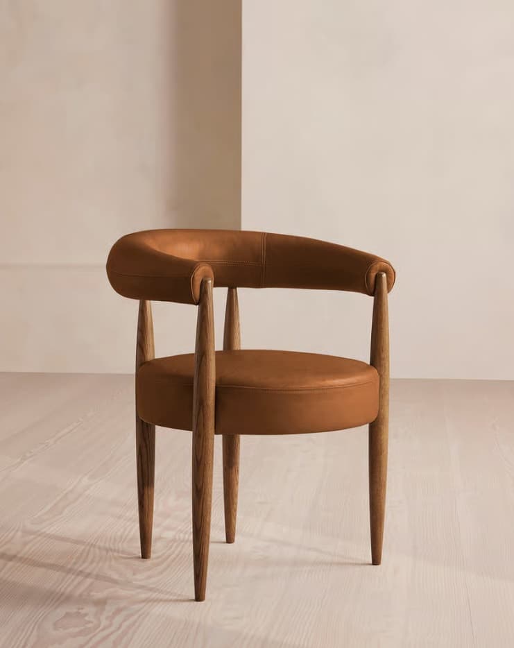 A modern armchair featuring a dark wood frame and saddle leather upholstery. The chair has a curved, continuous backrest that flows into integrated armrests. The seat is a circular, padded cushion. It stands on four slender, tapered wooden legs. — 3D model