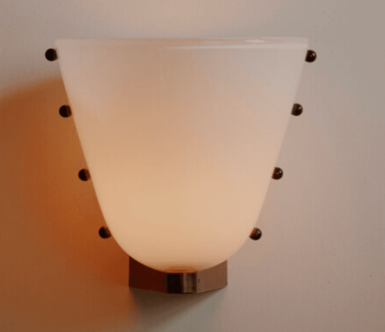 A wall-mounted lighting fixture, commonly known as a wall sconce, featuring an opaque white or frosted glass shade. The shade has a tapered, bowl-like form that opens upwards. It is secured to the wall via a metallic backplate and held in place by multiple small, dark, spherical studs or fixings positioned along the vertical edges of the glass shade. The fixture emits a soft, diffused light, characteristic of frosted glass diffusers. — 3D model