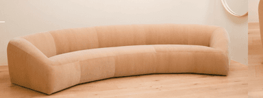 A contemporary, generously curved upholstered sofa in a light, neutral beige tone. The sofa features a continuous, rounded form with no visible legs, giving it a low-profile and substantial presence. The backrest and seat are seamlessly integrated, suggesting a cohesive, single-piece construction. The upholstery appears to be a textured fabric, possibly boucle or a fine-grained velvet, with subtle vertical seams segmenting the back and seat portions. Its design is minimalist, focusing on organic curves and comfort. — 3D model
