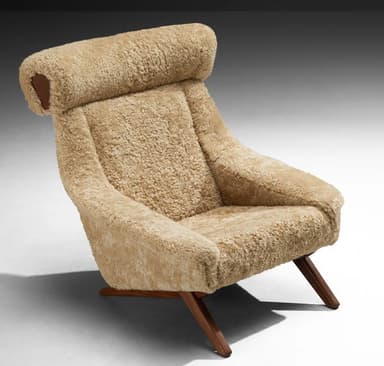 A high-backed armchair with a distinctive sloped silhouette. The chair is upholstered entirely in a textured, light brown sheepskin or shearling material, giving it a plush and organic appearance. The seat and back cushions are integrated into the overall form. The base consists of dark wood legs with a visible grain, angled outwards for stability. The headrest area is pronounced and rolls over the top of the backrest. — 3D model