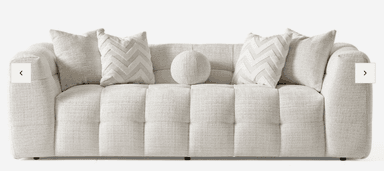 A long, low-profile contemporary sofa upholstered in a light beige, textured fabric. It features deep, square tufting across the seat, backrest, and integrated armrests, creating a plush and inviting appearance. Several decorative pillows in matching and complementary patterns are arranged on the sofa. — 3D model