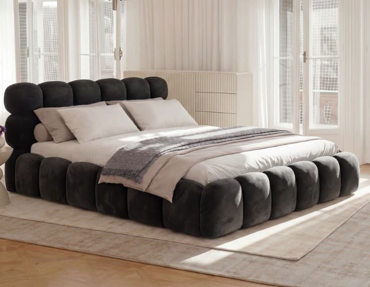 A contemporary upholstered bed frame with a distinctive rounded, modular headboard and a low-profile base. The headboard is composed of stacked, curved segments that create a soft, organic silhouette. The bed is dressed with white linens, a light grey blanket, and several pillows, including standard rectangular pillows and a cylindrical bolster pillow. — 3D model