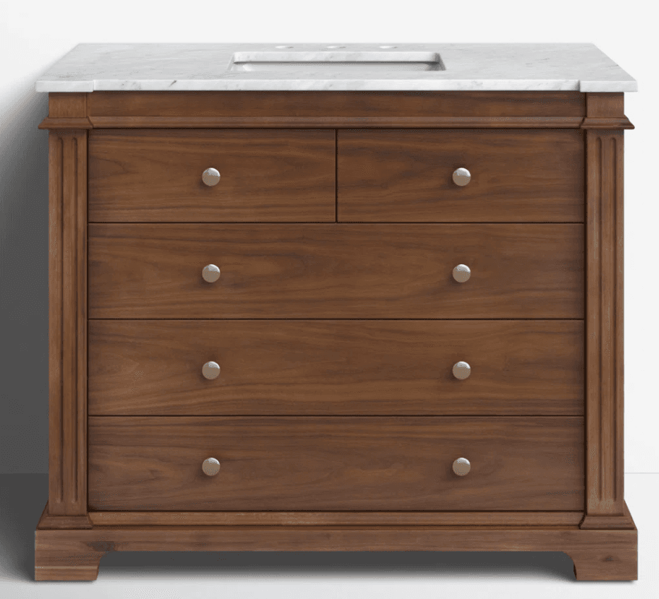 A traditional style bathroom vanity constructed from rich brown wood, featuring a white marble countertop with an integrated sink basin. The vanity has a rectangular form with fluted pilaster details on the side stiles and rests on a plinth base. It is equipped with multiple drawers of varying sizes, each fitted with round, satin brass-toned knobs. — 3D model