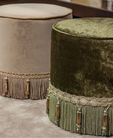 A cylindrical ottoman upholstered in a green velvet fabric. It features a decorative trim around the top edge and a prominent fringe with wooden bead accents at the base. — 3D model