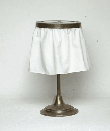 A table lamp featuring an antiqued brass finish pedestal base, a slender brass stem, and a distinctive draped white fabric shade. The shade is suspended from a circular brass frame with an integrated cross-patterned grille at the top, allowing for light diffusion. The white fabric drapes softly, creating a unique silhouette. — 3D model