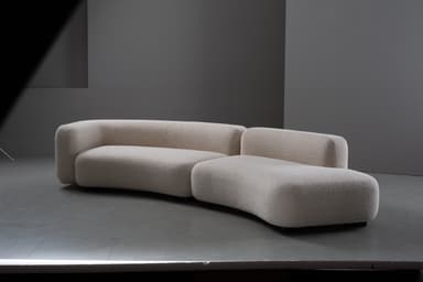 A two-piece modular sofa featuring a distinct curved silhouette and upholstered in a light-colored, textured boucle fabric. The design is contemporary with rounded edges and a low profile, comprising a left arm section and a right armless chaise-like section that together form a gentle curve. The sofa sits close to the floor on concealed or minimalist feet. — 3D model