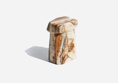 A small, monolithic piece of furniture carved from a single block of variegated marble, featuring a flat, octagonal-like top surface supported by a thick, sculpted base with a distinct 'T' or inverted 'L' profile and subtly faceted sides. The stone displays natural white, cream, and beige tones with prominent brown, rust, and grey veining. — 3D model