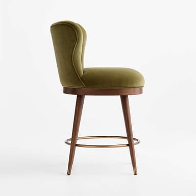 A stylish bar stool featuring an olive green velvet upholstered seat and a distinctive scalloped, channel-tufted backrest. It stands on four angled wooden legs, connected by a circular brass footrest, and finished with brass ferrules on the feet. The design blends luxurious materials with mid-century modern influences. — 3D model
