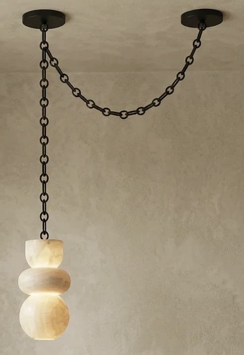 A contemporary pendant light featuring a sculptural shade crafted from translucent alabaster, suspended by a black metal chain system. The shade is composed of three stacked, organic, rounded forms, illuminated from within to highlight the natural veining and luminous quality of the stone. The suspension system consists of a long chain dropping from one ceiling mount, connecting to the light fixture, and a shorter chain extending horizontally to a second ceiling mount, creating a distinctive draped effect. — 3D model