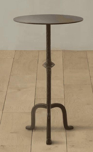 A tall, dark metal pedestal table featuring a round, flat top with a subtle hammered texture. The central column has a pronounced hammered finish and a decorative bulge midway down, transitioning into three elegantly curved legs that provide a stable, tripod base. The finish is a matte, dark bronze or antique iron. — 3D model