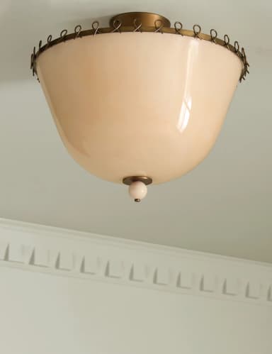 A semi-flush mount ceiling light featuring a domed, opaque off-white opaline glass shade. The shade is suspended from a brass ceiling canopy and encircled by a decorative band of small, looped brass elements at its upper edge. A small, spherical finial in a matching off-white tone protrudes from the bottom center of the glass shade, secured by a brass fitting. — 3D model
