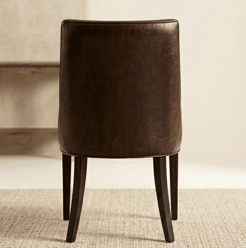 A dark brown dining chair viewed from the back. The chair features a curved, upholstered backrest and a seat base supported by four tapered wooden legs. The upholstery appears to be leather or a similar material with a matte finish. The legs are dark and slender, angled slightly outwards. — 3D model