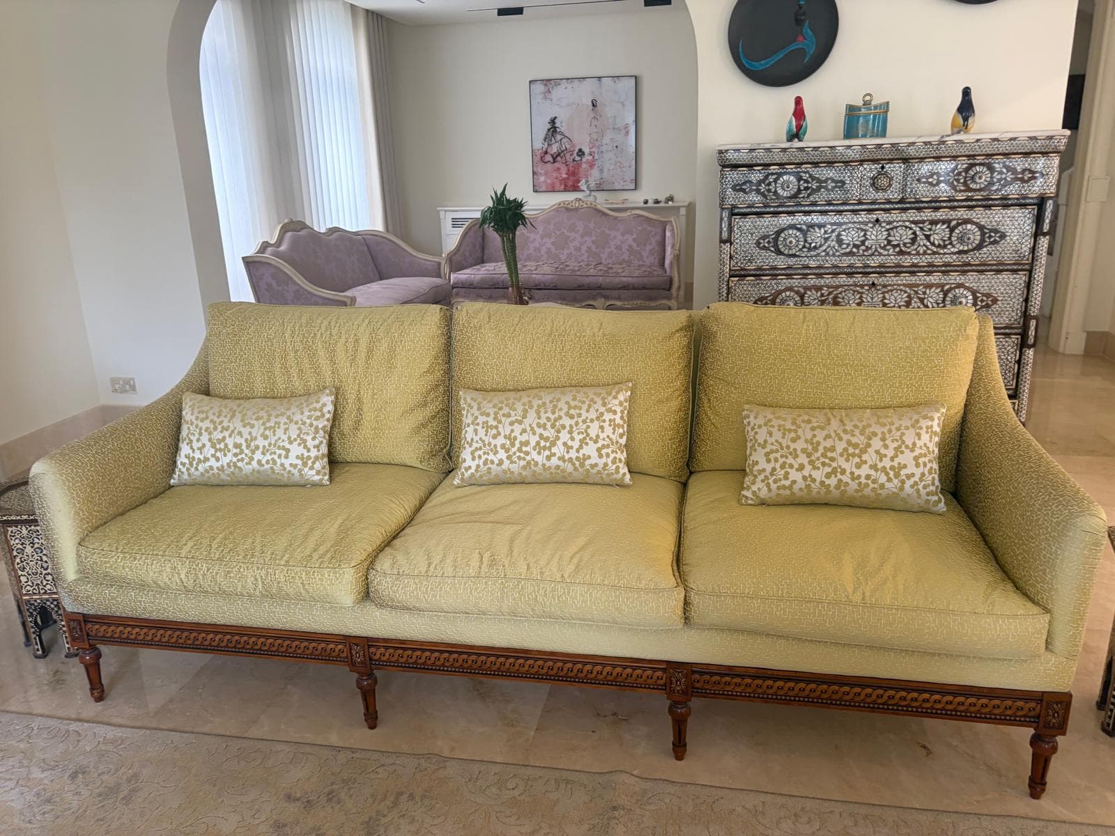A three-seater sofa featuring a light yellow-green textured upholstery, a dark wood frame with ornate carvings along the base and legs, and rolled arms. It includes three seat cushions, three back cushions, and three smaller decorative throw pillows with a lighter, mottled pattern. — 3D model