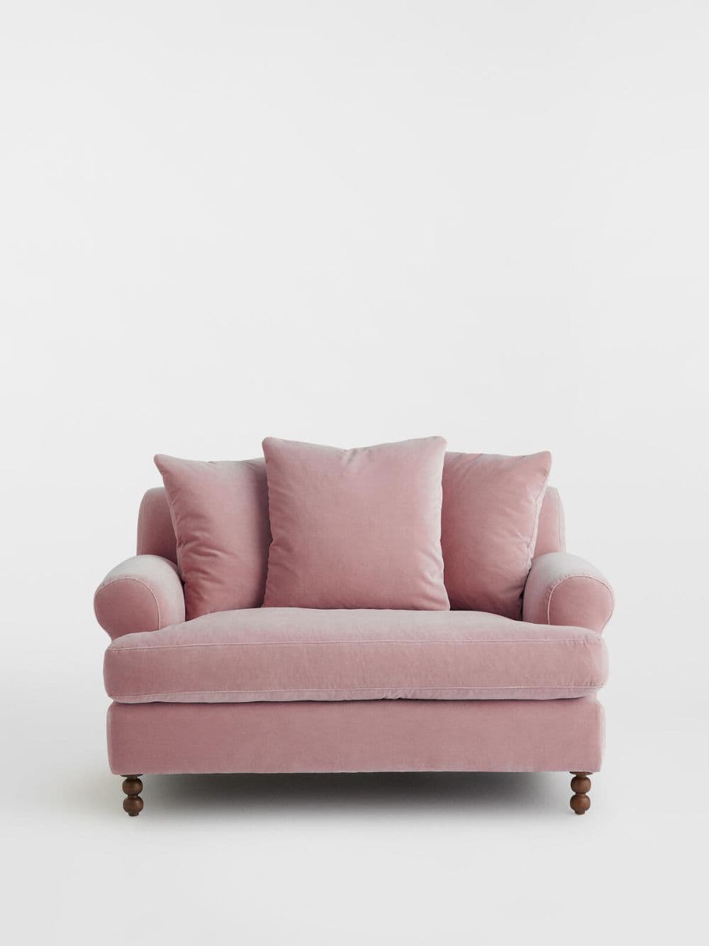 A plush pink sofa with rolled arms, featuring three throw pillows and dark wooden, turned legs. The upholstery appears to be a soft velvet fabric. The sofa has a compact, two-seater design. — 3D model