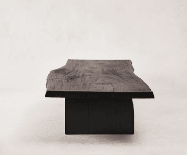A low, rectangular coffee table with a live-edge top and thick, blocky legs. The tabletop has an irregular, natural edge on one side, and the surface displays visible wood grain. The legs are positioned inset from the ends of the tabletop. — 3D model