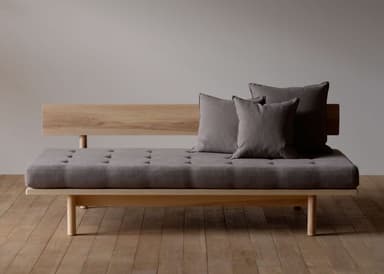 A modern daybed featuring a light-toned wooden frame and a grey upholstered cushion. The frame includes a simple slatted backrest on one side and stands on cylindrical wooden legs. The main cushion is tufted, and two matching throw pillows are included. — 3D model