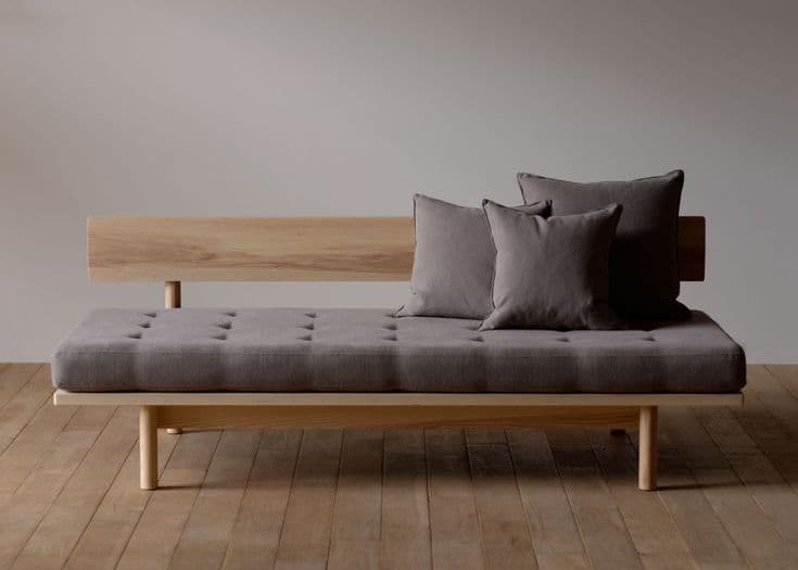 A modern daybed featuring a light-toned wooden frame and a grey upholstered cushion. The frame includes a simple slatted backrest on one side and stands on cylindrical wooden legs. The main cushion is tufted, and two matching throw pillows are included. — 3D model