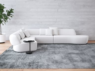 A large, light-colored modular sectional sofa composed of multiple interconnected units. It features a distinctive curved armrest section, several straight seating modules with integrated backrests, and a extended chaise lounge-style end unit. The sofa is upholstered in a textured, light-toned fabric and includes several loose throw pillows for added comfort. A small, black C-shaped side table with a round base is positioned next to the curved armrest section. — 3D model