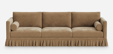 A long, rectangular three-seater sofa featuring a full-length skirted base and track arms. The sofa is upholstered in a soft, possibly velvet or chenille, fabric of a light brown or beige color. It has three separate back cushions and three seat cushions, providing defined seating areas. Each arm is accompanied by a cylindrical bolster cushion. The skirt conceals the legs and adds a traditional or transitional aesthetic. — 3D model