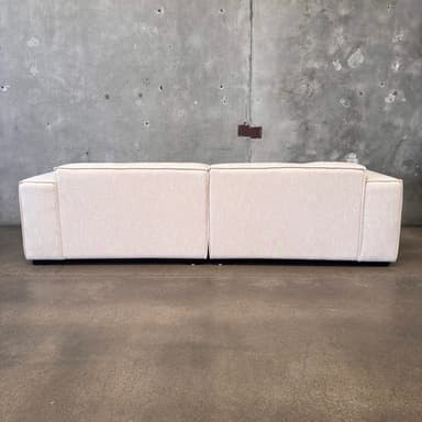 A modern, two-seater sofa with a low profile and blocky arms. The sofa is upholstered in a light-colored, textured fabric and features contrasting dark piping along the edges of the cushions and frame. The base appears to be a dark, solid plinth. It is positioned against a rough, concrete wall with visible imperfections and mounted fixtures. — 3D model