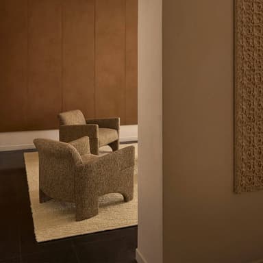 A modern, upholstered armchair featuring a distinctive sculptural form, wide integrated arms, and a sturdy base with curved cut-outs. It is covered in a textured, brown fabric, presenting a contemporary aesthetic. — 3D model