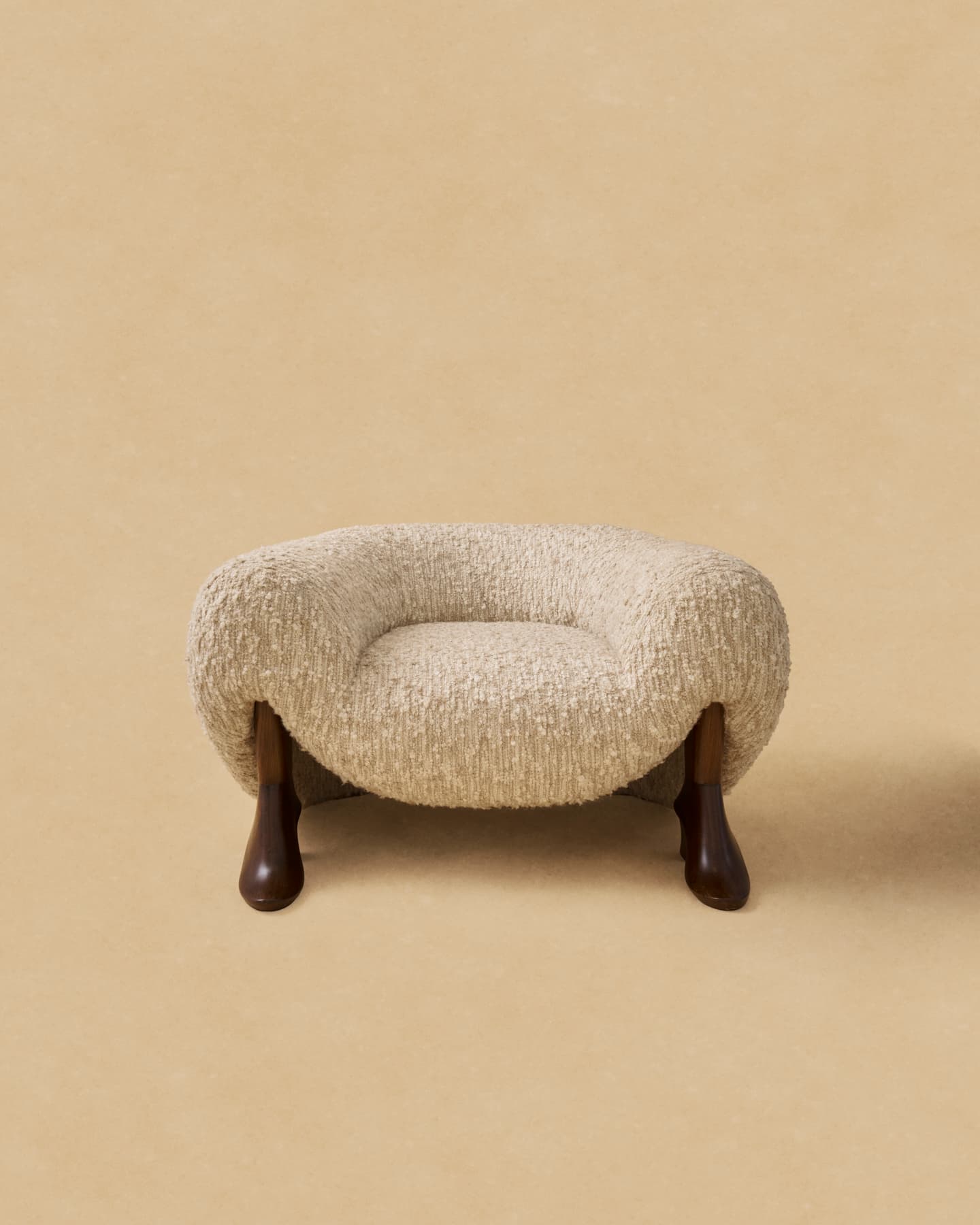 An armless lounge chair featuring a fully upholstered body in a cream-colored, textured bouclé fabric, supported by four dark, organically shaped wooden legs. The chair has a low profile and a rounded, inviting silhouette. — 3D model