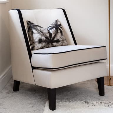 A contemporary upholstered accent armchair featuring off-white or cream fabric, distinguished by black contrasting piping along the edges of the frame and cushions. It stands on four tapered black legs and includes a decorative throw pillow with a black and grey abstract pattern. — 3D model