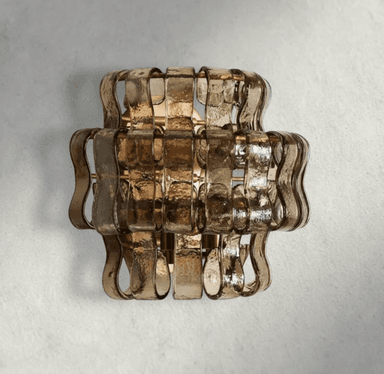 A contemporary wall sconce featuring multiple horizontal bands of textured, smoked or amber-toned glass, arranged in a layered, undulating pattern. The glass panels are secured by an underlying metallic frame, likely brass-finished, which is partially visible between the glass sections. The fixture is designed for vertical mounting and provides diffused, warm illumination. — 3D model