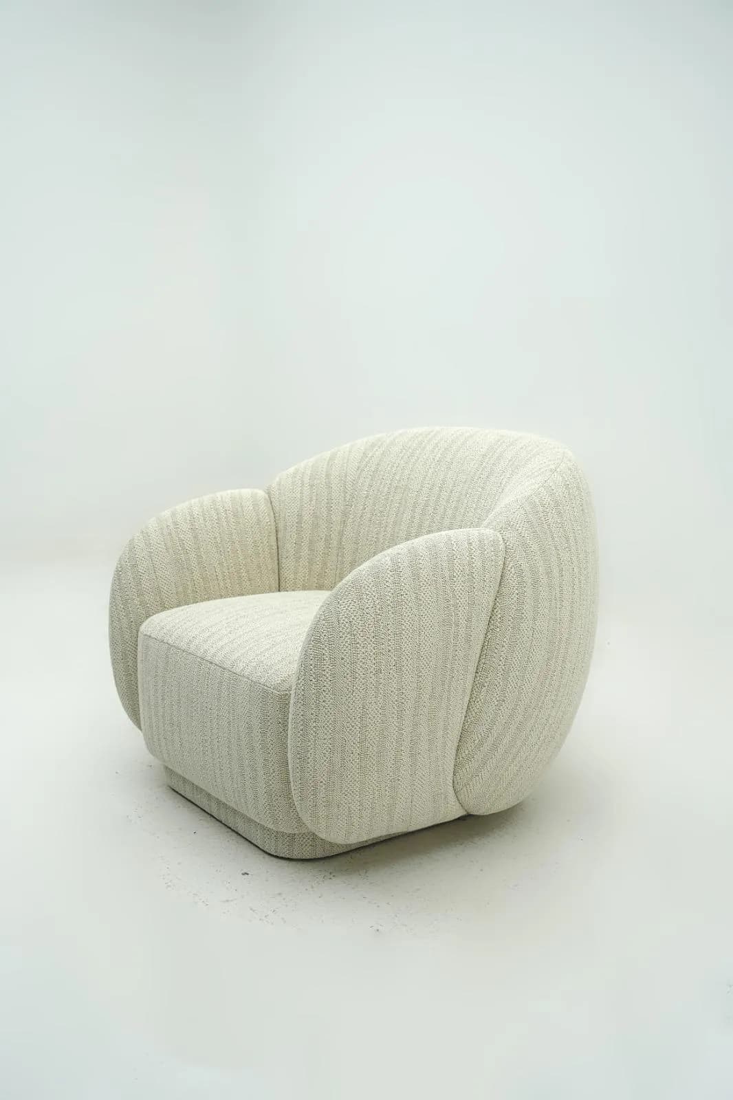 A modern, fully upholstered lounge chair featuring a rounded, sculptural silhouette. The chair has wide, integrated arms and a low back that curve around the seat, all covered in a light-toned, textured fabric with subtle vertical ribbing. The base is concealed, giving it a grounded appearance. — 3D model